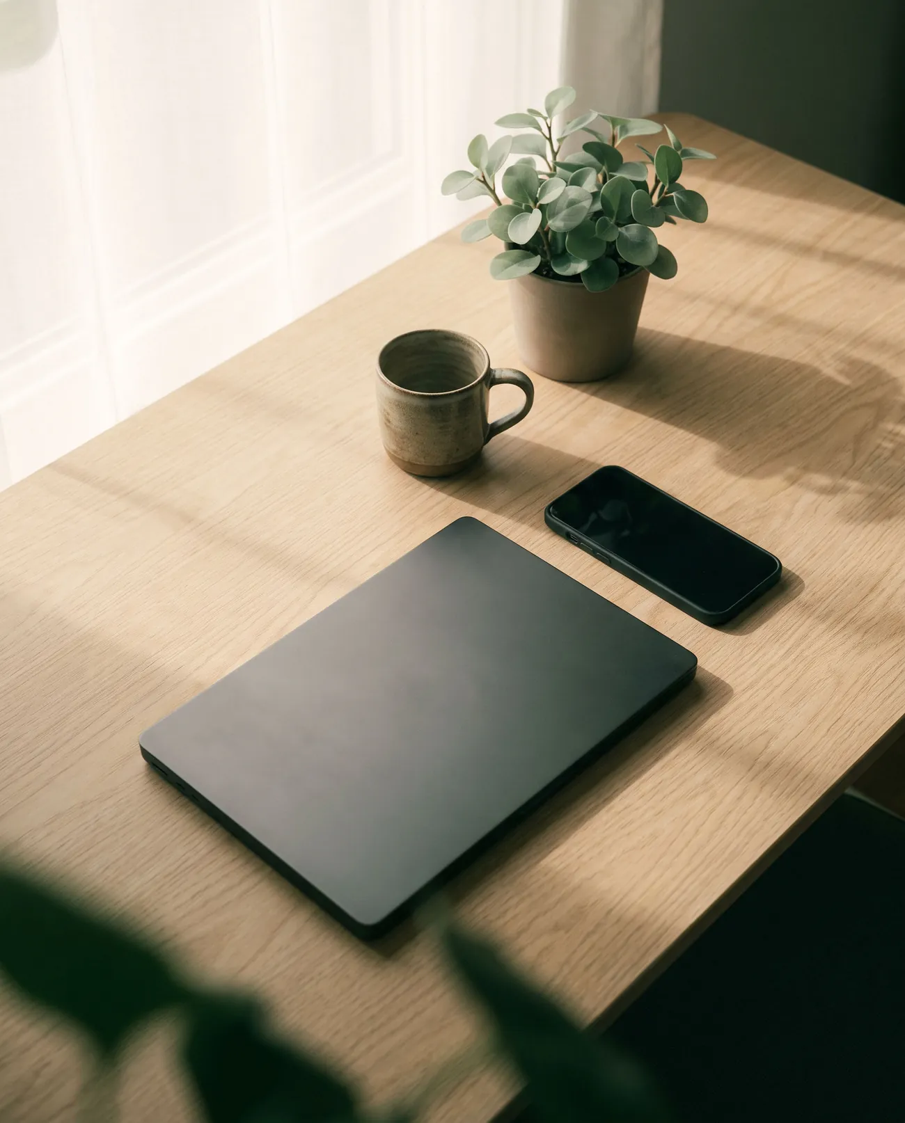 A calm, ordered oak desk with a closed laptop, a phone, a mug and a small plant.